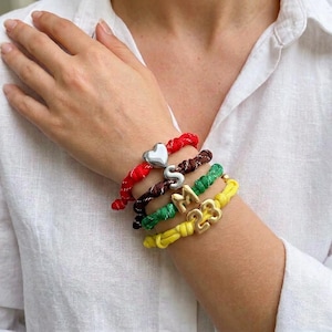 May include: A collection of colorful braided bracelets with silver and gold charms. The bracelets are red, brown, green, and yellow. Charms include a heart, the letter "S", the letters "M", and the numbers "23".