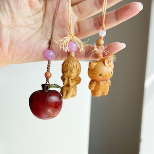 May include: Three wooden charms suspended on strings. The charms feature a red apple, a small angel, and a cartoon cat. Each charm has a decorative bead above it. The charms are held up by a hand.