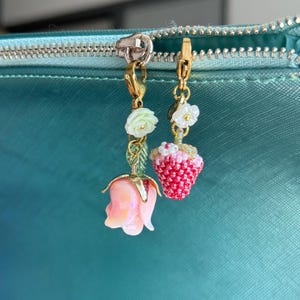 May include: Two gold-coloured charm pendants. One features a pink rose, the other a red and white beaded strawberry. Both charms have white flower and green leaf accents. The charms are hanging from a teal zip.