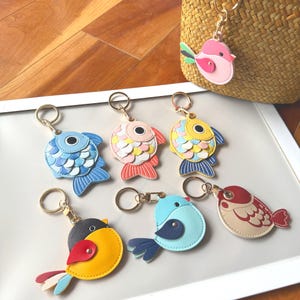 May include: A selection of vibrant fish and bird keyrings. The keyrings display a range of colours, such as blue, pink, yellow, and red, with gold-toned key rings. The fish keyrings have scales, and the bird keyrings have wings and tail feathers.