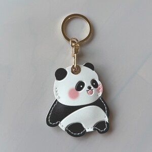 May include: A panda bear keychain with a gold-colored ring and clasp. The panda has a white body, black patches, and pink cheeks. The keychain is made of a leather-like material.