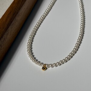 Peut inclure: Un collier de perles d&eacute;licat avec un charm dor&eacute;. Le collier est compos&eacute; de petites perles blanches rondes. Le charm dor&eacute; est une petite perle ronde. Le collier est pr&eacute;sent&eacute; sur une surface blanche.