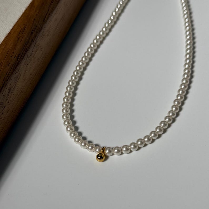 Peut inclure: Un collier de perles d&eacute;licat avec un charm dor&eacute;. Le collier est compos&eacute; de petites perles blanches rondes. Le charm dor&eacute; est une petite perle ronde. Le collier est pr&eacute;sent&eacute; sur une surface blanche.