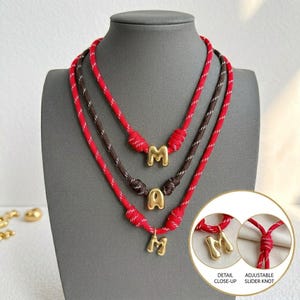 Puede incluir: Tres collares con cordones trenzados rojos y marrones con dijes de letras dorados. Los collares incluyen nudos deslizantes ajustables. Los dijes tienen forma de letra M. El recuadro muestra un primer plano detallado y un nudo deslizante ajustable.