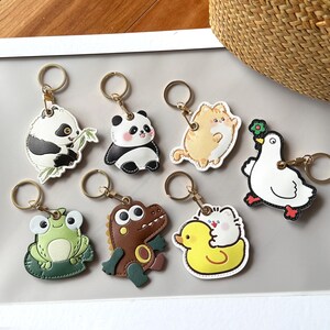 May include: A collection of colorful animal-shaped keychains. The keychains feature designs of a panda, cat, duck, frog, dinosaur, and a bird. Each keychain has a gold-colored ring for attaching to keys or bags.