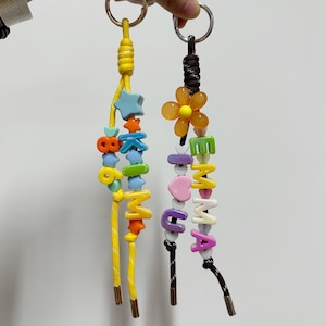 Puede incluir: Dos llaveros con cuentas de colores que deletrean palabras. Uno es amarillo con cuentas azules, naranjas y verdes. El otro es marrón con una flor y cuentas que deletrean "MAMMA". Ambos tienen llaveros de metal.