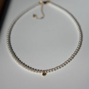 Peut inclure: D&eacute;licat collier de perles avec un petit pendentif dor&eacute;. Le collier est compos&eacute; de petites perles rondes et d'un charm dor&eacute;. Le fermoir est &eacute;galement dor&eacute;. Le collier est pr&eacute;sent&eacute; sur une surface blanche.
