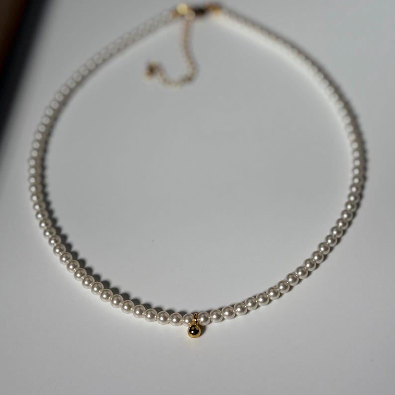 Peut inclure: D&eacute;licat collier de perles avec un petit pendentif dor&eacute;. Le collier est compos&eacute; de petites perles rondes et d'un charm dor&eacute;. Le fermoir est &eacute;galement dor&eacute;. Le collier est pr&eacute;sent&eacute; sur une surface blanche.