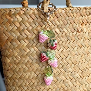 Pu&ograve; includere: Un ciondolo a forma di fragola con perline, con bacche rosa e rosse, foglie verdi e una catena dorata. Il ciondolo &egrave; attaccato a una borsa di paglia intrecciata con una chiusura dorata.