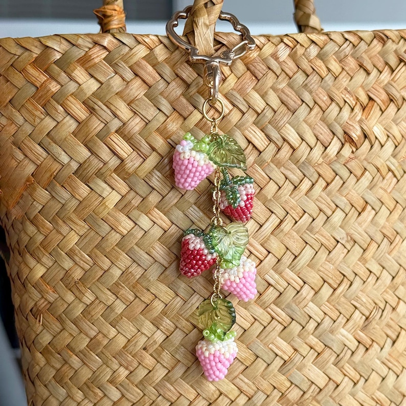 Pu&ograve; includere: Un ciondolo a forma di fragola con perline, con bacche rosa e rosse, foglie verdi e una catena dorata. Il ciondolo &egrave; attaccato a una borsa di paglia intrecciata con una chiusura dorata.