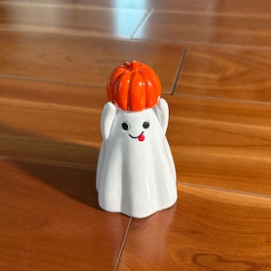 Puede incluir: Una figura de fantasma blanco con una cara sonriente y una lengua roja. El fantasma lleva un sombrero de calabaza naranja. La figura est&aacute; sobre una superficie de madera.