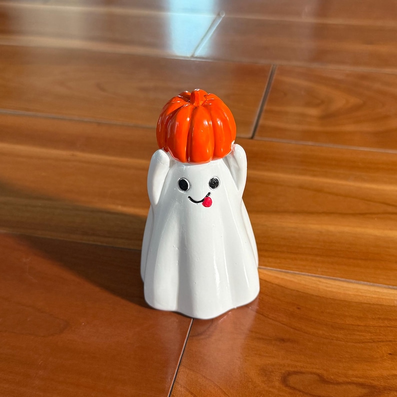 Puede incluir: Una figura de fantasma blanco con una cara sonriente y una lengua roja. El fantasma lleva un sombrero de calabaza naranja. La figura est&aacute; sobre una superficie de madera.