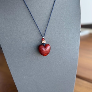 以下が含まれることがあります： 濃い赤色の木製ハートのペンダントネックレス。 ネックレスは濃い青色のコードと、ハートの上に小さな装飾的なビーズクラスターが付いています。 ハートペンダントの幅は約3.8cmです。