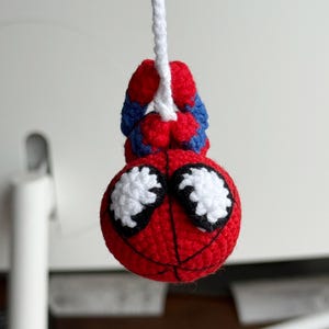 May include: A handmade crochet Spider-Man figure, featuring a red head with white eyes and black outlines, and a red and blue body. The figure is suspended by a white string, showcasing the character's iconic colors and design.