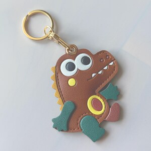 May include: A brown leather dinosaur keychain with a gold-colored keyring and clasp. The dinosaur has large white-rimmed eyes, a yellow belly, and green feet. The keychain is a fun accessory.