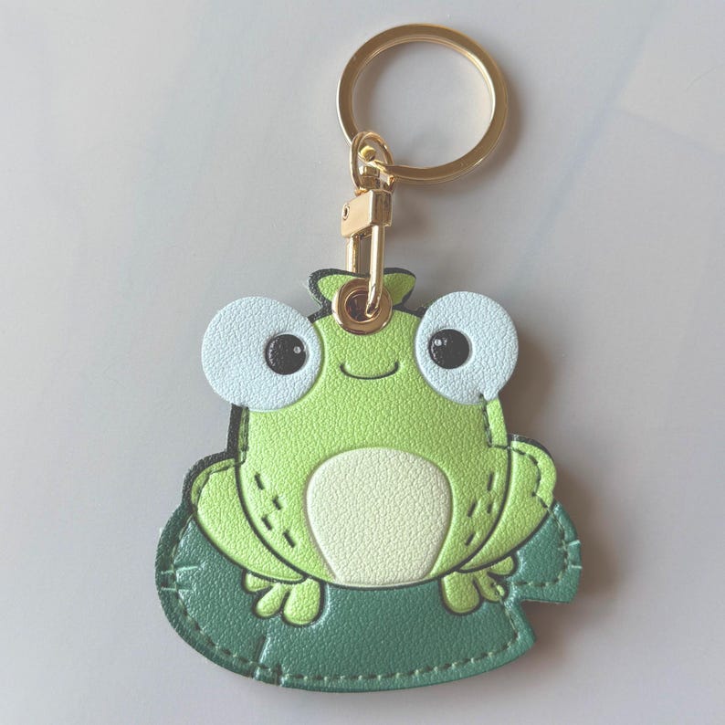 May include: A green frog-shaped keychain with a gold-colored keyring. The frog has large, light blue eyes and a cream-colored belly. It is sitting on a green lily pad. The keychain is made of leather or a similar material.