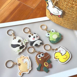 May include: A collection of animal-themed keychains. The keychains feature designs of a panda, frog, cat, alligator, duck, and a goose. Each keychain has a gold-colored ring for attaching to keys or bags. The keychains are made of leather-like material.