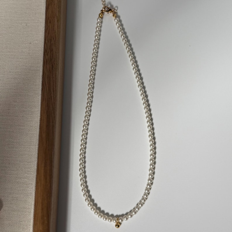 Peut inclure: Un collier de perles d&eacute;licat avec un fermoir en or et un petit pendentif en or. Le collier est compos&eacute; de nombreuses petites perles blanches rondes enfil&eacute;es ensemble. Les accents dor&eacute;s contrastent subtilement.