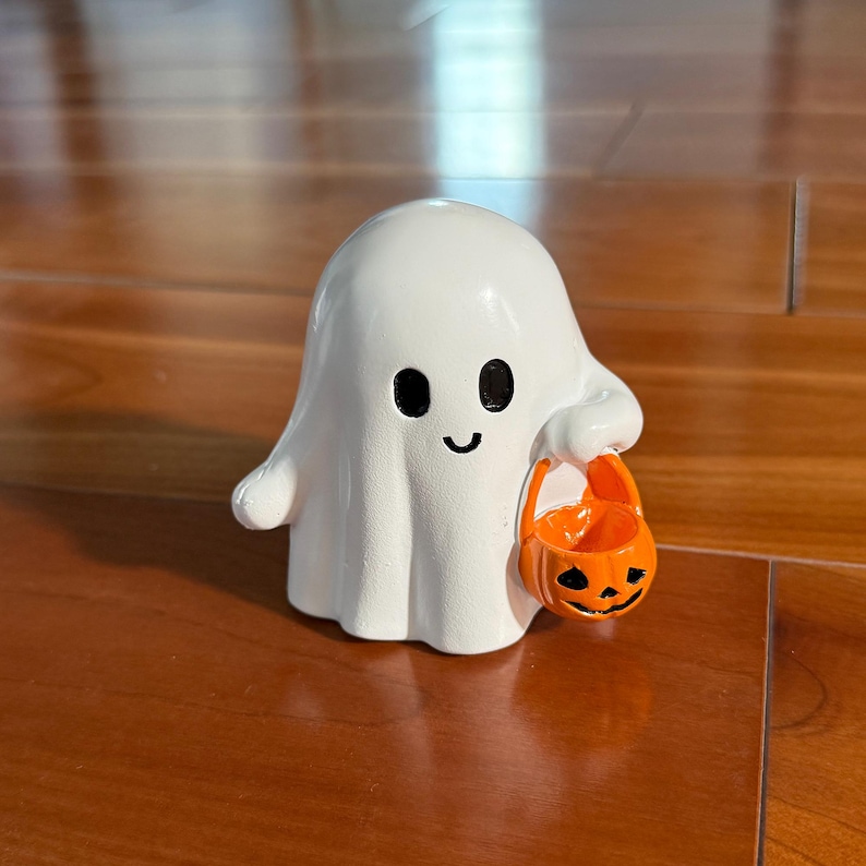 Puede incluir: Una figura de fantasma blanco con ojos negros y boca curva, sosteniendo una cesta de calabaza naranja. La decoraci&oacute;n tem&aacute;tica de Halloween est&aacute; sobre una superficie de madera.