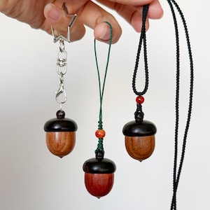 Puede incluir: Tres colgantes en forma de bellota con tapas de color marrón oscuro y cuerpos de madera natural. Uno está unido a un llavero plateado en forma de estrella, otro a un cordón verde con cuentas rojas y el tercero a un cordón negro.