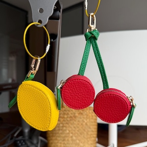 May include: Three small, round coin purses in the shape of a lemon and cherries. The lemon purse is yellow, and the cherry purses are red. Each purse has a gold zipper and a green stem. They hang from gold keychains.