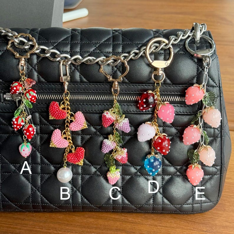 Pu&ograve; includere: Cinque portachiavi decorativi con disegni di fragole. Ogni portachiavi presenta un ciondolo a forma di fragola di stile diverso, tra cui varianti rosse, rosa e blu, con finiture color oro. I portachiavi sono attaccati a una borsa nera trapuntata.