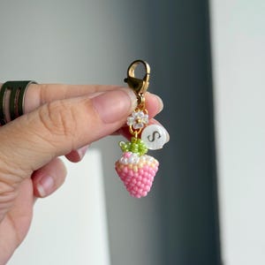 May include: A beaded strawberry charm with a gold-coloured clasp. The strawberry is pink and white, with green leaves and a small white flower. A white circular bead with the letter "S" is also attached.