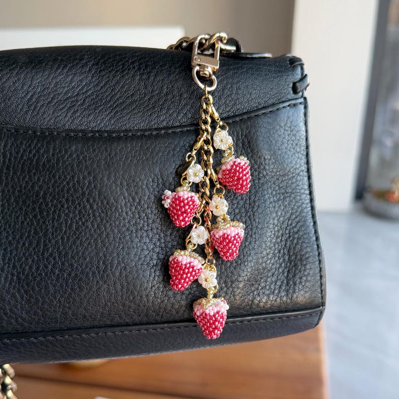 Pu&ograve; includere: Un ciondolo a forma di fragola con perline con catena e chiusura color oro, attaccato a una borsa in pelle nera. Il ciondolo presenta pi&ugrave; fragole rosse e bianche con accenti floreali bianchi, creando un accessorio stravagante.