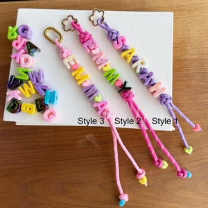 May include: Three personalized keychains with names spelled out in colorful letter beads. Each keychain features a different color scheme and design, with the names "SARAH", "EMMA", and "MOMMY". The keychains are attached to a gold-colored clasp.