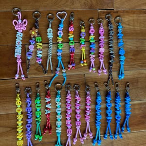 May include: A collection of twenty personalised keychains featuring colourful plastic letter beads, various shaped charms like hearts, flowers, and stars, and braided cords attached to metal lobster claw clasps. Each keychain displays a unique name.
