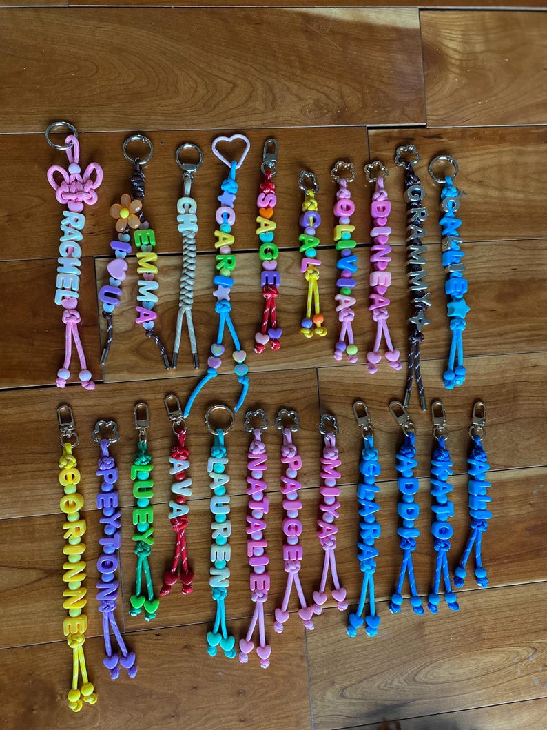May include: A collection of twenty personalised keychains featuring colourful plastic letter beads, various shaped charms like hearts, flowers, and stars, and braided cords attached to metal lobster claw clasps. Each keychain displays a unique name.