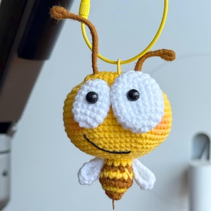 May include: A handmade, crocheted bee with a yellow body, white eyes, and brown antennae. The bee has a smiling face and small white wings. The bee is hanging from a yellow cord.