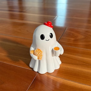 Puede incluir: Una figura de fantasma blanco con un sombrero rojo, sosteniendo un cruas&aacute;n y una taza de caf&eacute;. El fantasma tiene ojos negros y una boca sonriente. La figura est&aacute; sobre una superficie de madera.