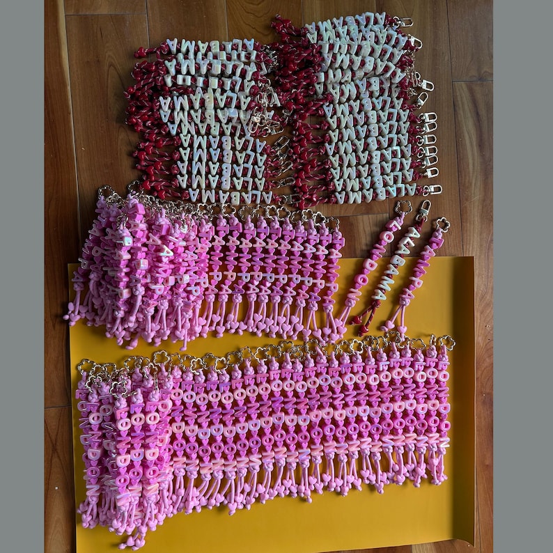 May include: A collection of beaded keychains featuring white letter beads spelling ALABAMA on red cords, and pink letter beads spelling TAMPA or LONDON on pink cords, all attached to gold-toned metal clasps with flower-shaped accents.