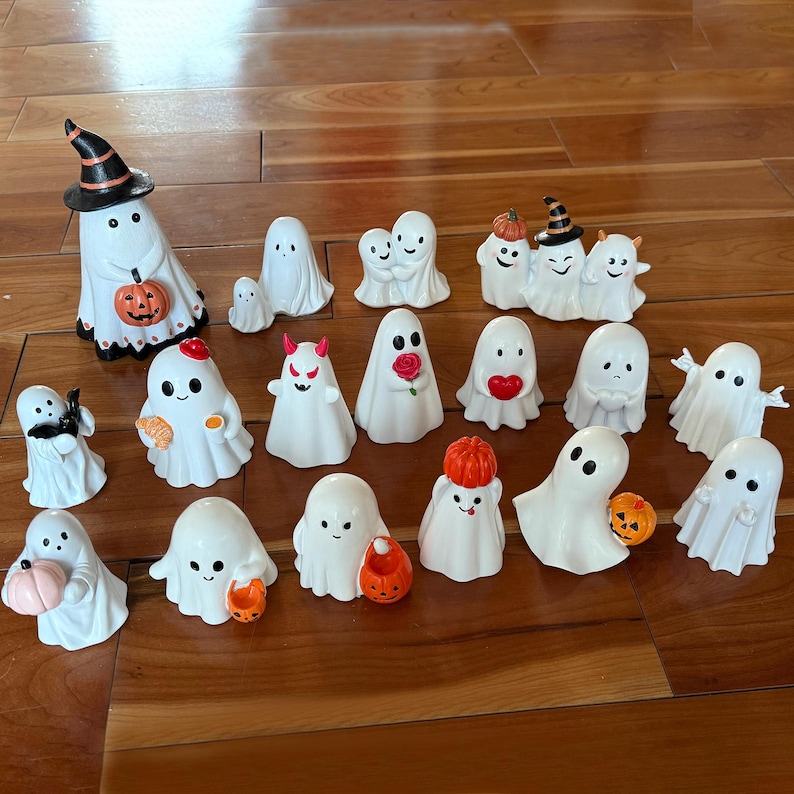 Puede incluir: Una colecci&oacute;n de figuras de fantasmas de cer&aacute;mica blanca con varios accesorios tem&aacute;ticos de Halloween. Algunos fantasmas llevan sombreros de bruja, sostienen calabazas o tienen cuernos de diablo. Las figuras est&aacute;n dispuestas sobre una superficie de madera.
