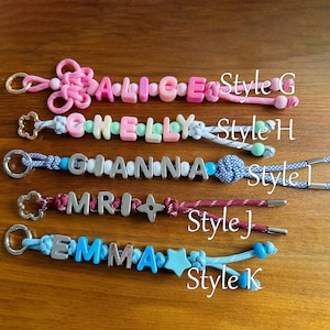 May include: Five personalised keychains with metal rings. Style G features pink cord and beads spelling ALICE. Style H has white cord and pastel beads spelling CHELLY. Style I uses blue patterned cord and silver letters spelling GIANNA. Style J has maroon cord and silver letters spelling MRI. Style K features light blue cord and blue letters spelling EMMA.
