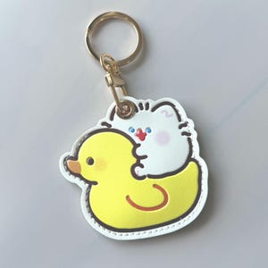 May include: A white cat hugging a yellow rubber ducky keychain. The keychain has a gold-colored ring and clasp. The cat has blue eyes and a red mouth. The duck has an orange beak and a red belly.