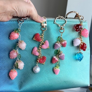 May include: Several beaded strawberry and heart charms hang from keychains. The charms are in shades of pink, red, and purple, with gold-colored chains and accents. The background is a teal-colored pouch.
