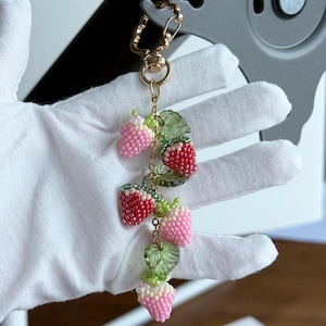 May include: A gold-coloured keyring with a beaded strawberry charm. The charm features multiple strawberries in pink and red, with green leaf details. The strawberries are made of small beads.