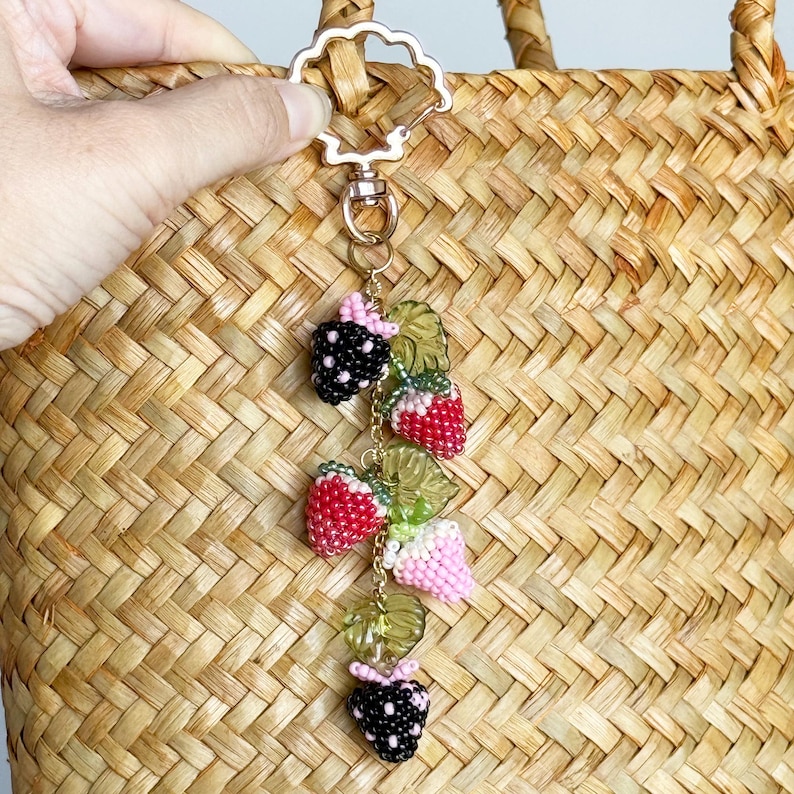Pu&ograve; includere: Un ciondolo a forma di frutta con perline con fragole e more. Il ciondolo &egrave; attaccato a una chiusura color oro. Il ciondolo &egrave; appeso davanti a una borsa di paglia intrecciata. Le fragole sono rosse e le more sono nere.