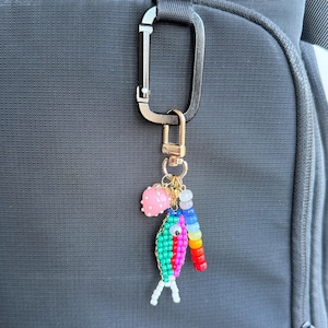May include: A colorful beaded keychain with a gold-tone clasp. The keychain features a beaded fish in green, pink, and red, a pink charm, and a rainbow-colored beaded accent. Attached to a black carabiner.