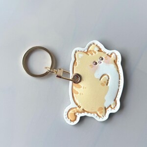 May include: A yellow and white cat-shaped keychain with a gold-colored keyring and clasp. The cat has orange stripes and a white belly. The keychain is made of a leather-like material.