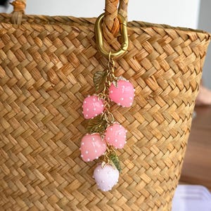 Pu&ograve; includere: Un ciondolo decorativo con un gruppo di ciondoli a forma di fragola rosa con accenti di perle bianche e foglie verdi. Il ciondolo &egrave; attaccato a una chiusura e una catena color oro, appeso a una borsa di paglia intrecciata.