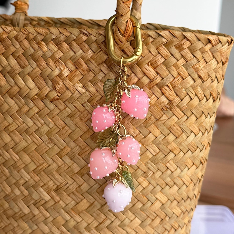 Pu&ograve; includere: Un ciondolo decorativo con un gruppo di ciondoli a forma di fragola rosa con accenti di perle bianche e foglie verdi. Il ciondolo &egrave; attaccato a una chiusura e una catena color oro, appeso a una borsa di paglia intrecciata.