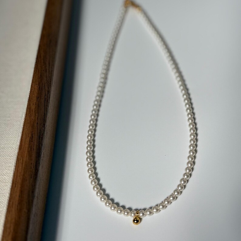 Peut inclure: Un collier de perles d&eacute;licat avec un petit pendentif en or. Le collier est compos&eacute; de petites perles blanches rondes. Le pendentif en or est une petite perle ronde. Le collier est pr&eacute;sent&eacute; sur une surface blanche.