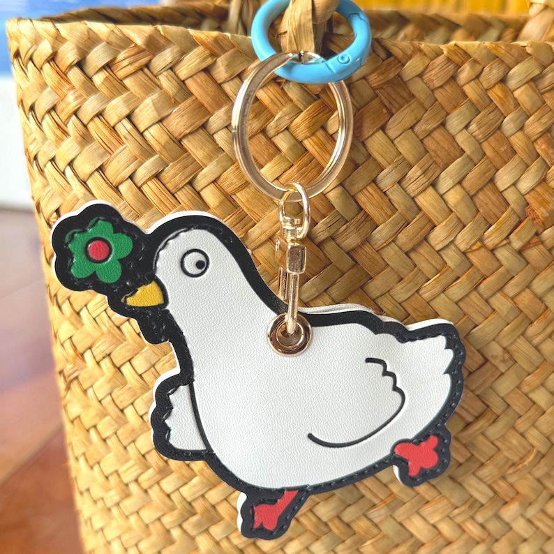 May include: A white dove keychain with a black outline, holding a green flower with a red center. The dove has a yellow beak and red feet. The keychain has a gold ring and a light blue clasp, attached to a woven basket.