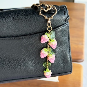 Pu&ograve; includere: Un ciondolo a forma di fragola con perline con bacche rosa e bianche e foglie verdi &egrave; appeso a una catena e a una chiusura color oro. Il ciondolo &egrave; attaccato a una borsa in pelle nera. Il ciondolo &egrave; un accessorio decorativo.
