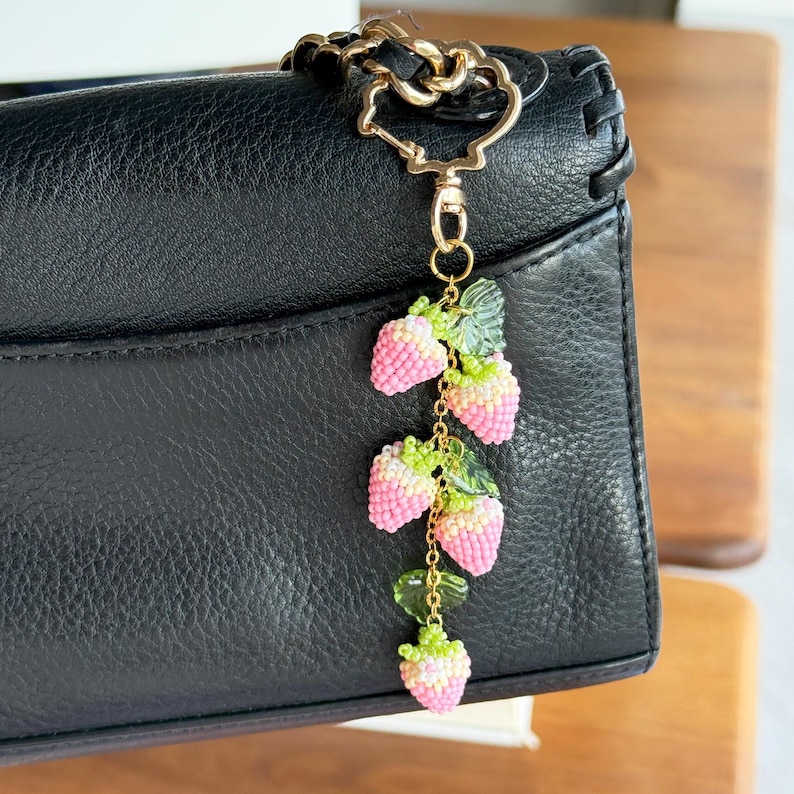 Pu&ograve; includere: Un ciondolo a forma di fragola con perline con bacche rosa e bianche e foglie verdi &egrave; appeso a una catena e a una chiusura color oro. Il ciondolo &egrave; attaccato a una borsa in pelle nera. Il ciondolo &egrave; un accessorio decorativo.