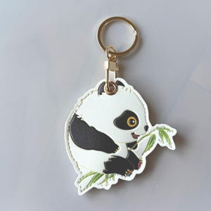 May include: A white and black panda bear keychain with a gold-colored keyring and clasp. The panda is eating bamboo leaves. The keychain is made of a leather-like material.
