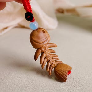 May include: A carved wooden fish skeleton pendant with a red cord. The pendant is light brown with detailed carving. A black button and a blue bead are on the cord above the pendant.